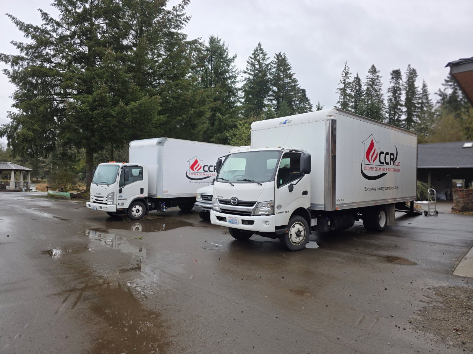 Two CCR box trucks parked side by side under tall evergreens